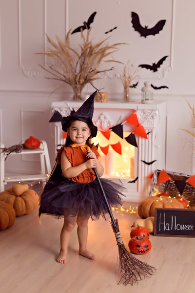 funny baby girl in halloween witch costume with pumpkin jack and broom indoors